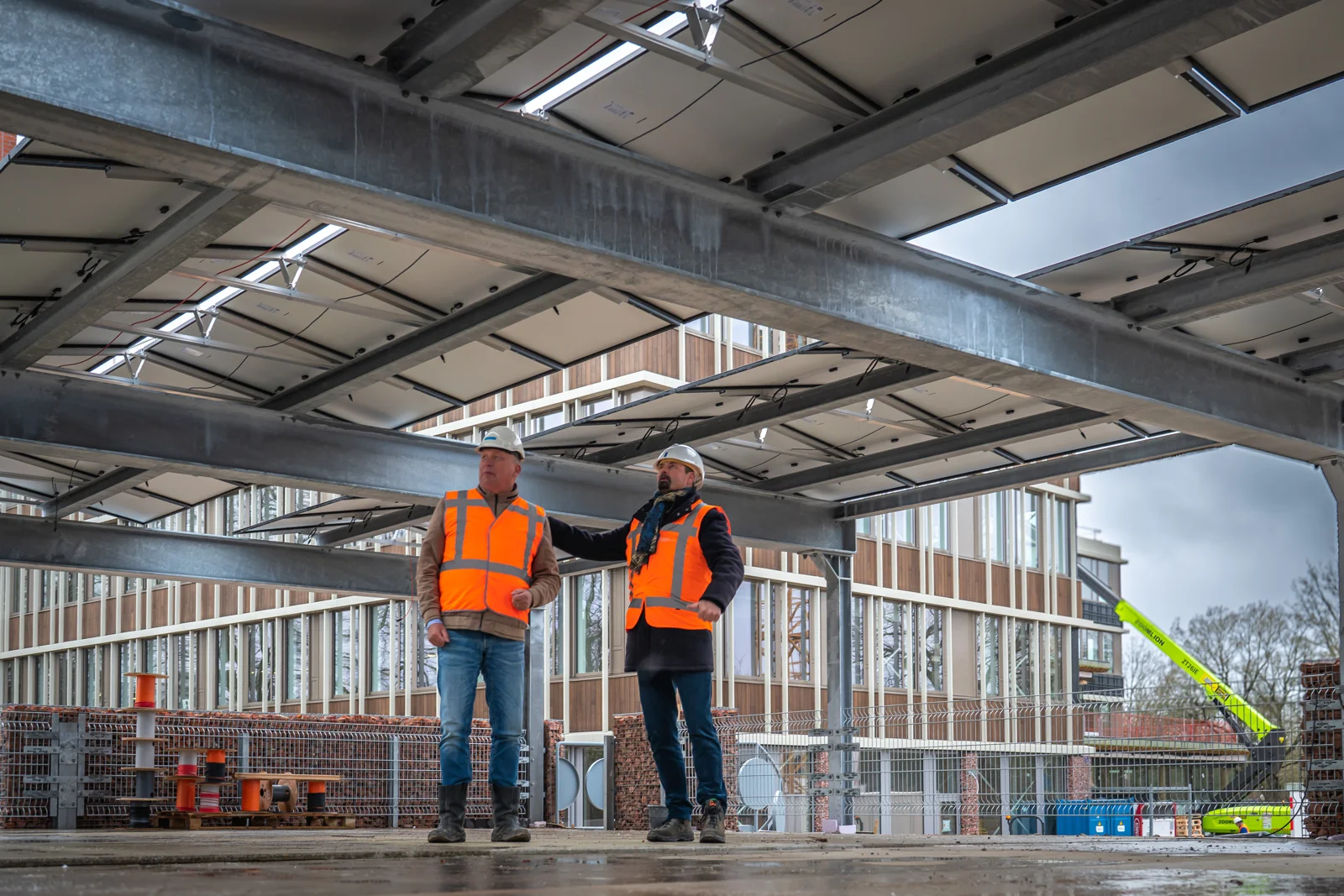 Twee mannen met bouwhelmen en oranje veiligheidsvesten onder een staalconstructie op een bouwplaats